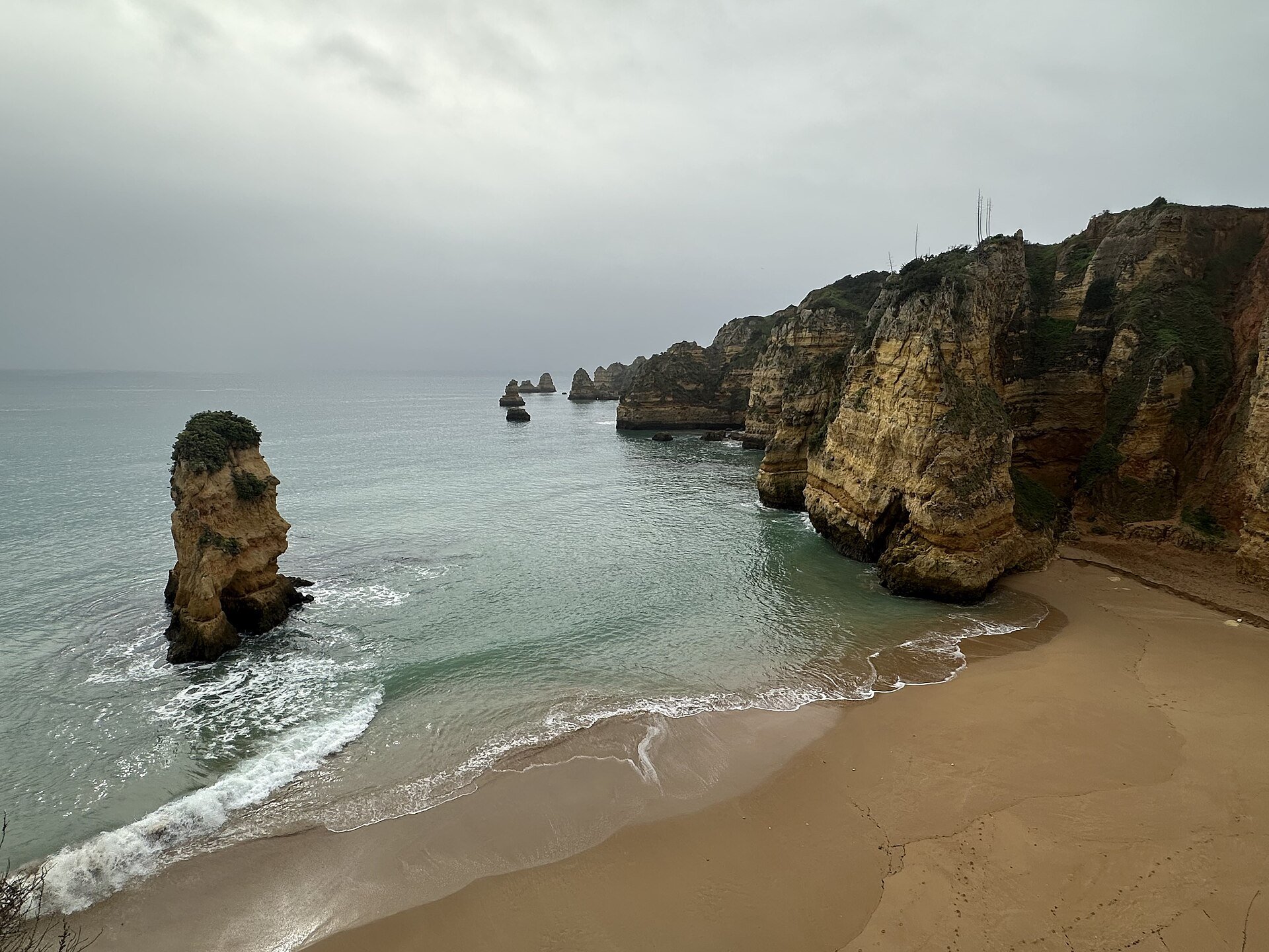 Sandy beach in Lagos, Faro, Algarve, Portugal, features prominent sea stacks and rugged cliffs. The overcast sky casts a subdued light over the scene. The sandstone formations rise dramatically from the clear, calm sea, highlighting natural